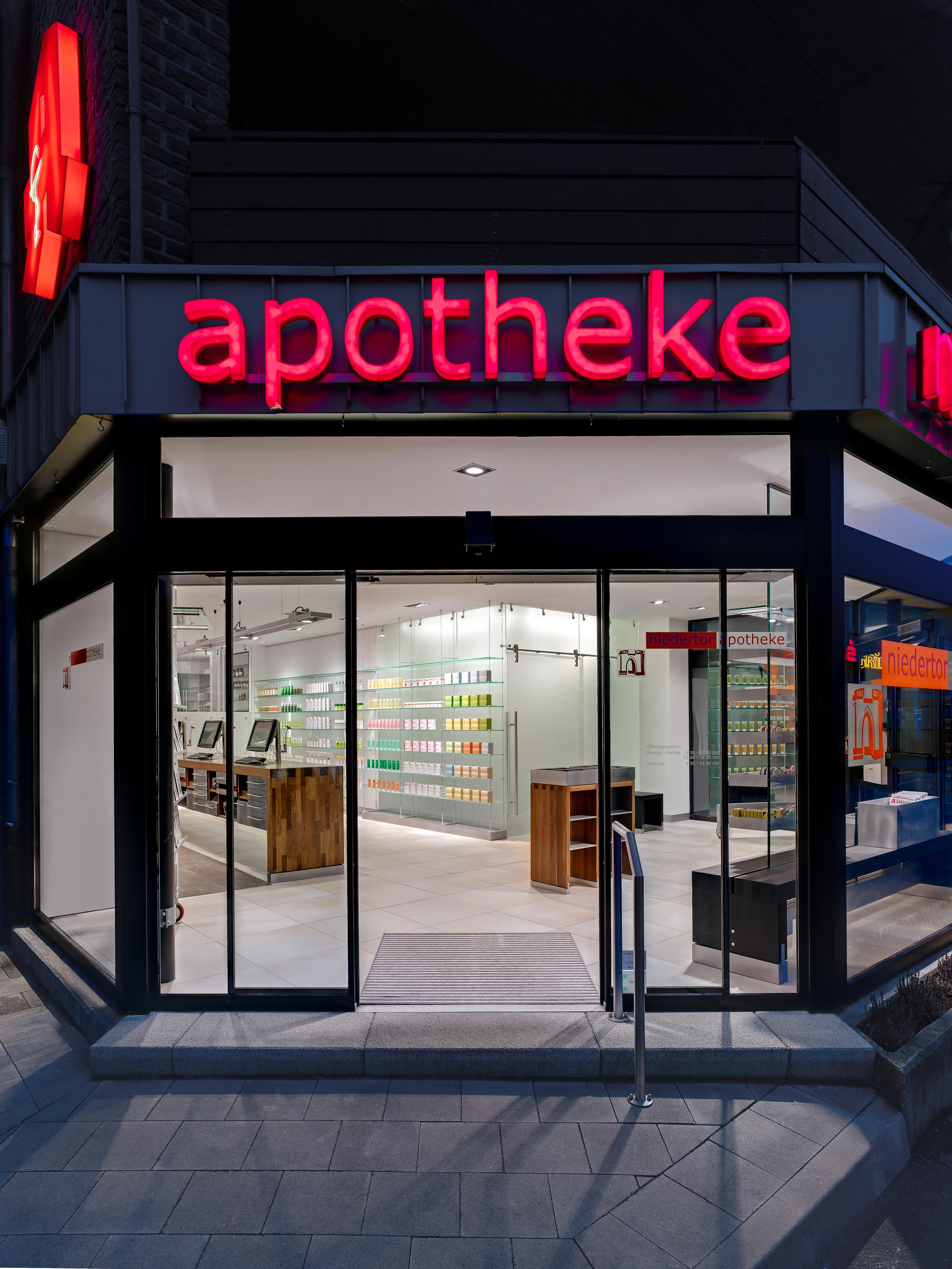 Abendansicht der Apotheke am Niedertor mit roter Leuchtschrift, Glasfassade und offenem Eingang
