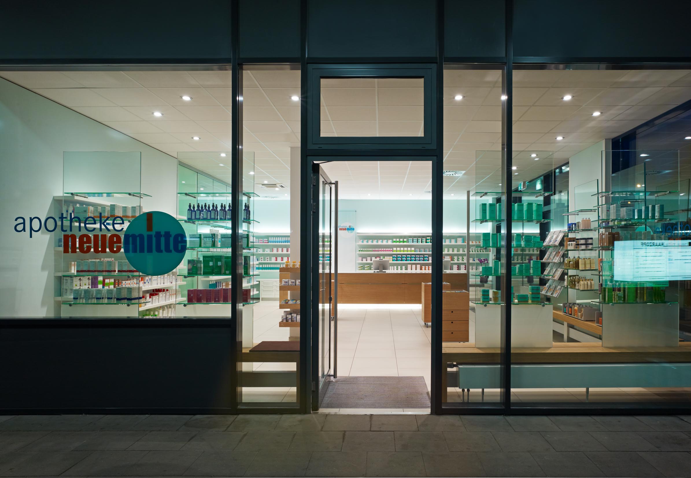 Abendansicht der Glasfassade mit Eingang und Einblick in die Apotheke Neue Mitte